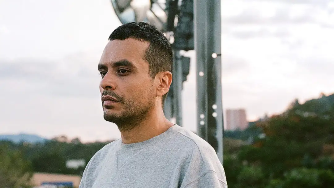MALA in a grey shirt stands in front of a building, looking towards the camera with a neutral expression.