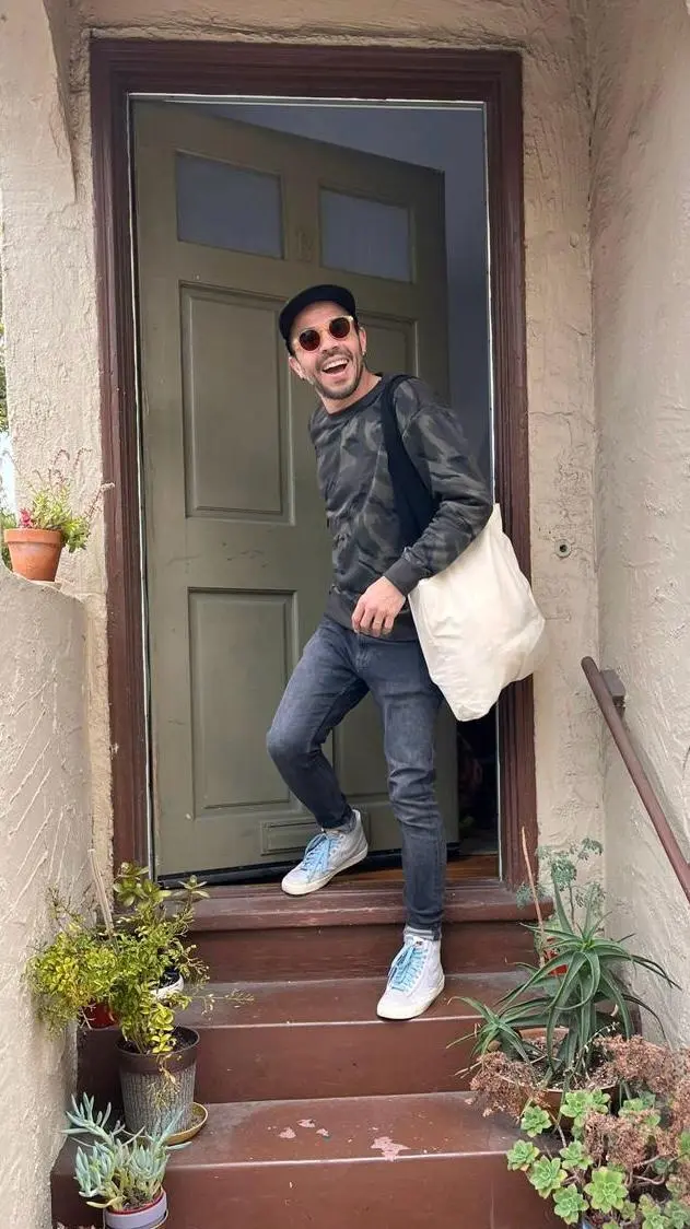 Qk wearing sunglasses and a hat stands on the steps of a house, smiling and looking towards the camera.