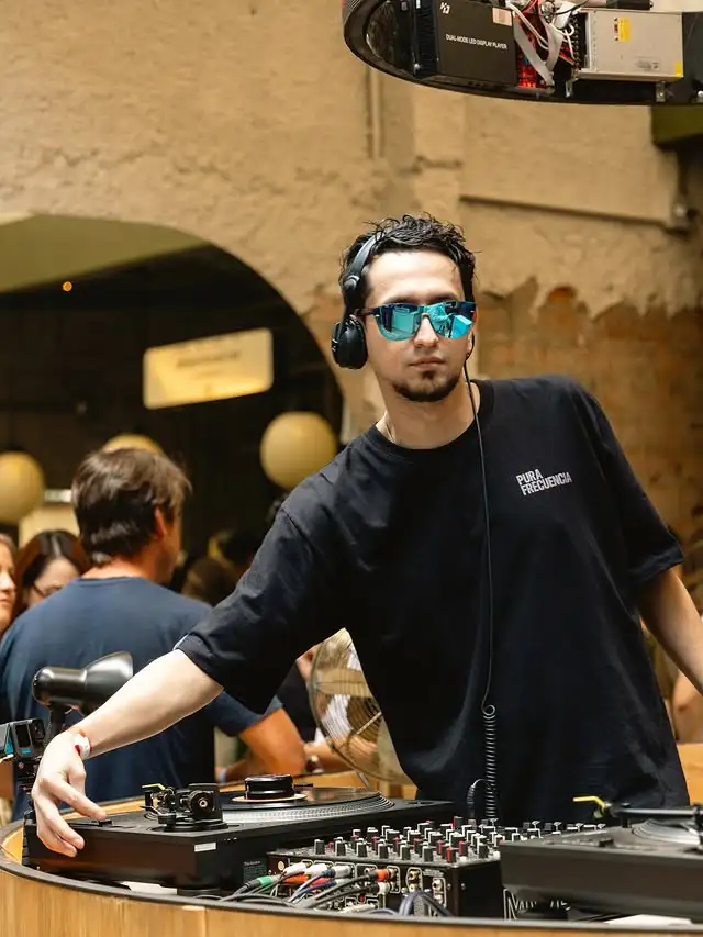 Sir Leaks wearing sunglasses mixes music at a lively party, surrounded by colorful lights and dancing guests.
