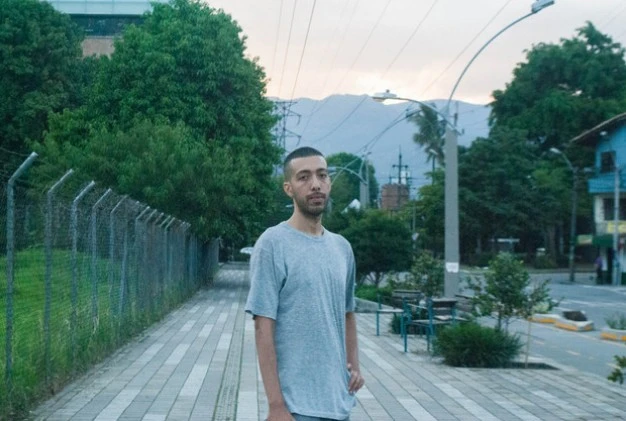 Therap stands on a sidewalk in front of a wooden fence, looking towards the camera with a neutral expression.
